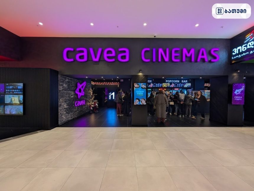 Purple neon 'cavea CINEMAS' sign above a dark theater entrance with people queuing and a small decorated tree to the left in a cinema lobby, hinting at a busy concession area.