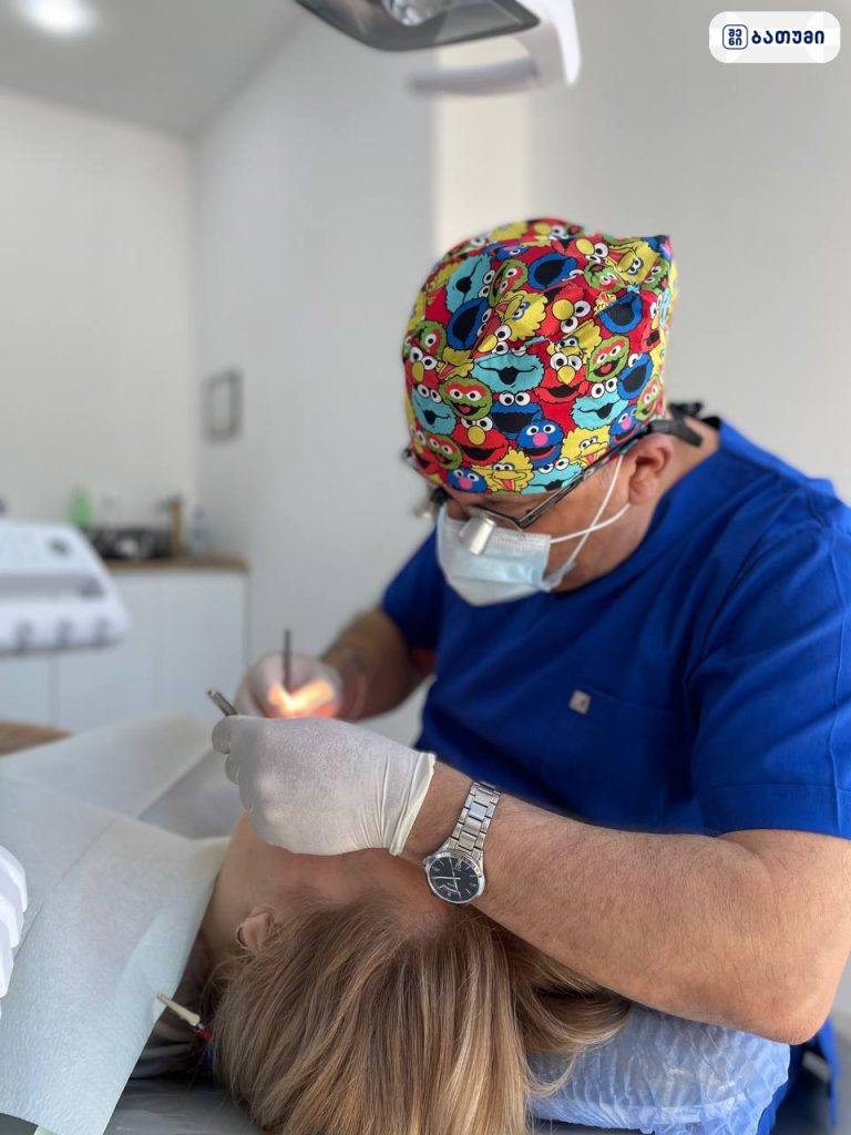 Dentist in a colorful cap and mask performs a dental procedure on a patient in a dental chair, using tools.