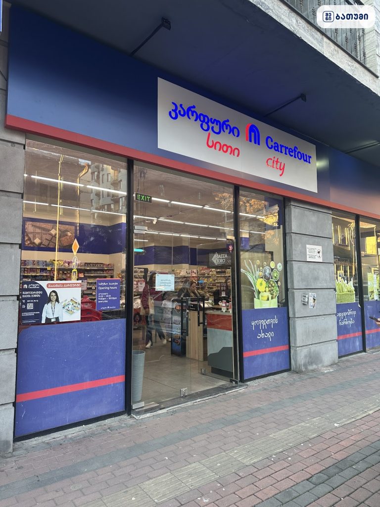 Exterior of Carrefour City store with blue-white signage and glass entrance; shoppers inside visible through the doors