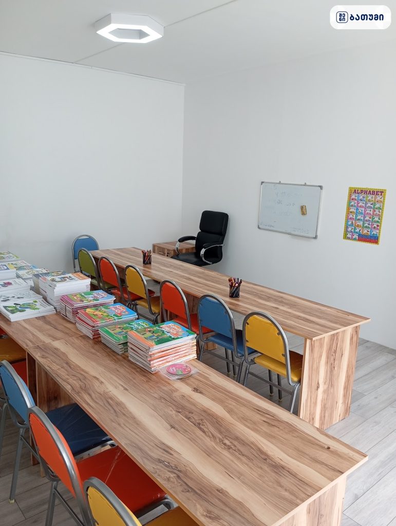 A bright classroom with long wooden tables lined with colorful child-sized chairs, stacks of books along the edge, and a black swivel chair at the far end near a whiteboard and alphabet poster on the wall
